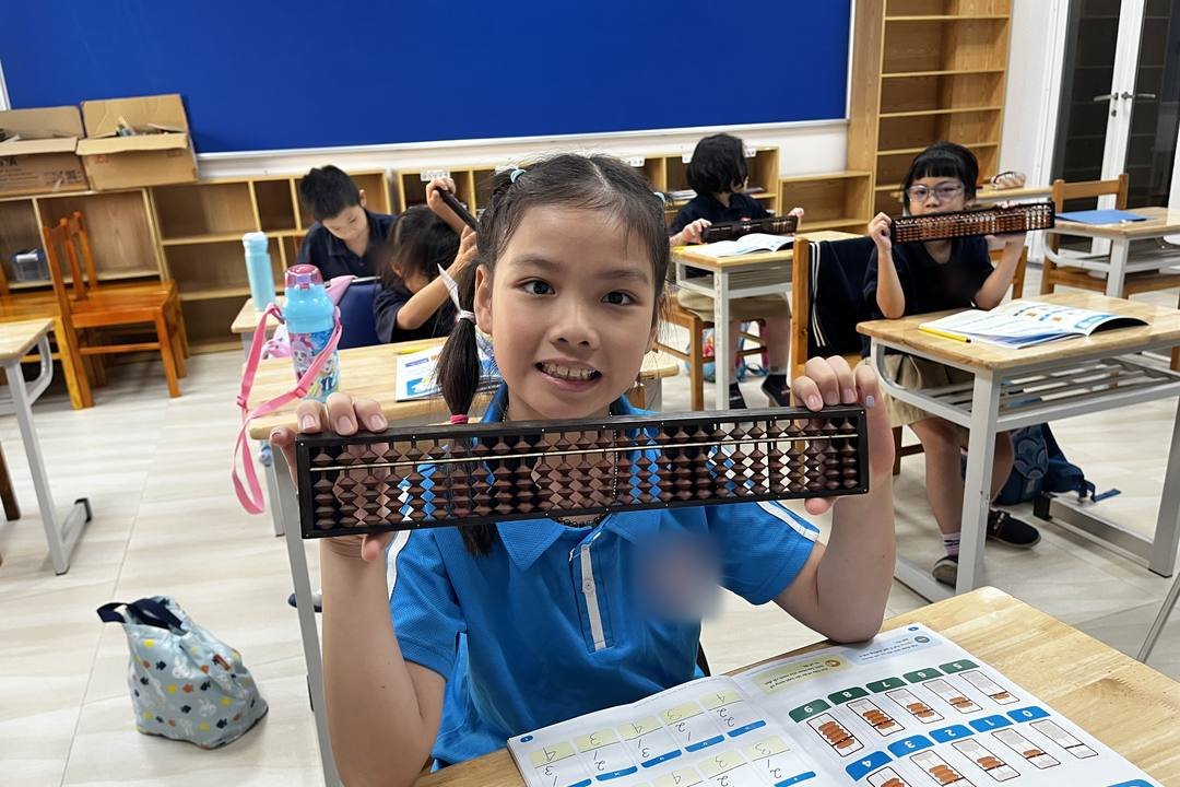 Bé gái mặc áo polo xanh dương cười tươi, hai tay giơ bàn tính Soroban; trên bàn là quyển sách Soroban đang mở, phía sau các bạn khác cũng đang luyện với bàn tính