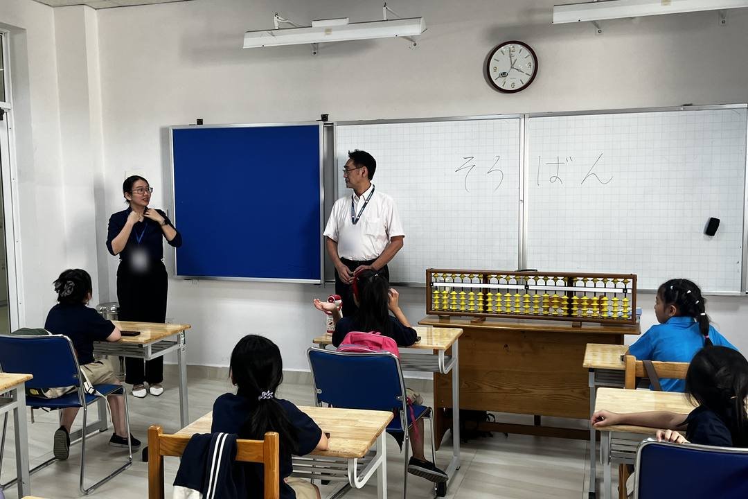 Đầu giờ học trong lớp câu lạc bộ Soroban: hai giáo viên đứng trước lớp, các bạn ngồi tại chỗ, trên bảng viết chữ “そろばん”