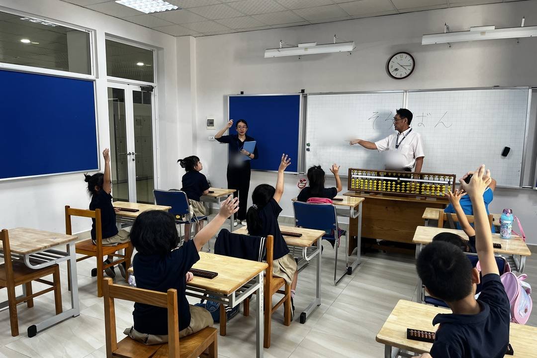 Học sinh tiểu học giơ tay trong giờ học Soroban; phía trước lớp có thầy cô, chiếc bàn tính lớn và chữ “soroban” viết bằng tiếng Nhật trên bảng