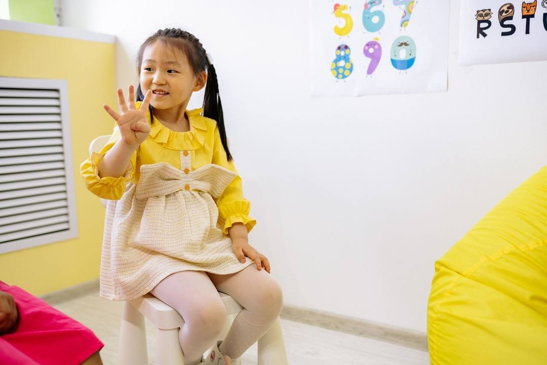 Bé gái mặc váy vàng ngồi trên ghế, mỉm cười giơ bàn tay lên, phía sau là tranh các chữ số trên tường