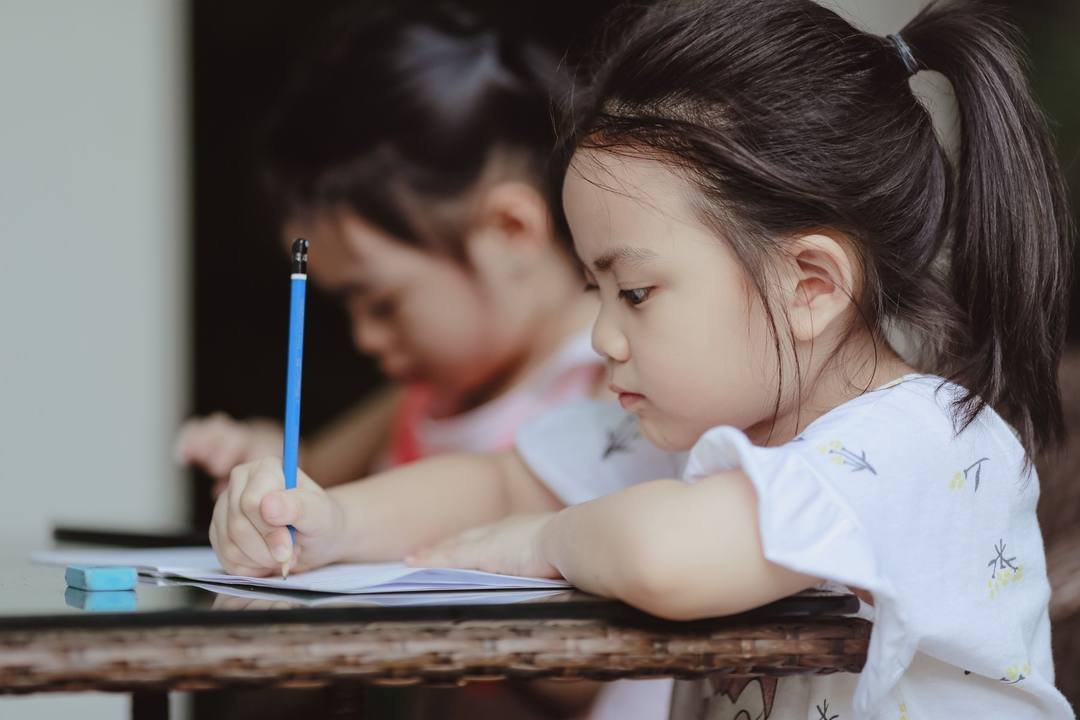 Hai bé gái ngồi bên bàn viết bài, bé phía trước buộc tóc đuôi ngựa, cầm bút chì xanh viết trên tờ giấy