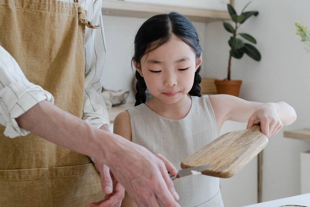 Bé gái cầm thớt gỗ trong bếp, đứng cạnh người lớn đeo tạp dề