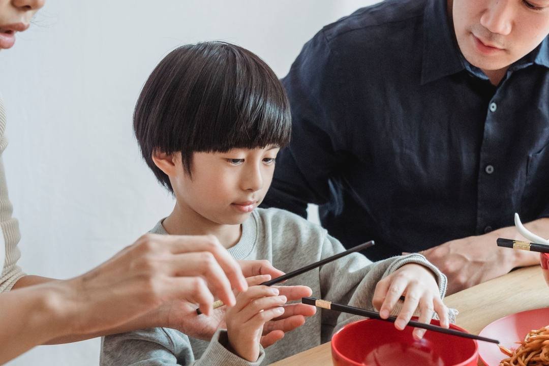 Bé trai ngồi giữa bố mẹ bên bàn ăn, tập cầm đũa; tay người lớn chỉ cho con cách cầm