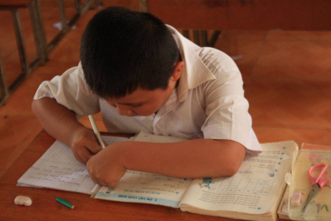 Cậu bé mặc áo sơ mi trắng cúi xuống viết vào vở, bên cạnh là quyển sách giáo khoa đang mở trên bàn gỗ
