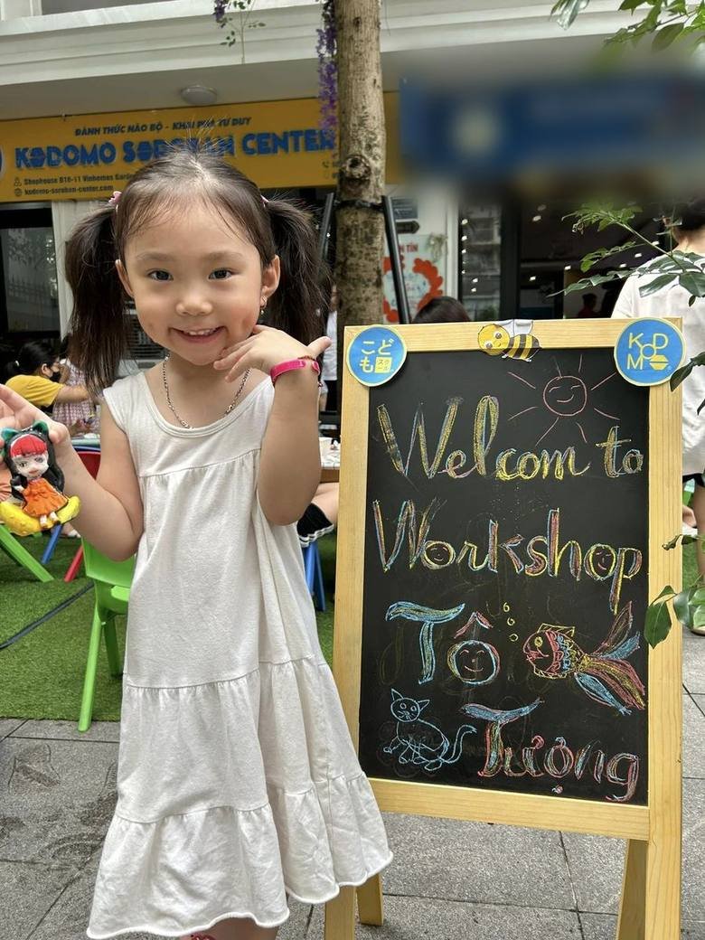 白いワンピースの女の子が「Welcome to Workshop Tô Tượng」の黒板の横に立っている