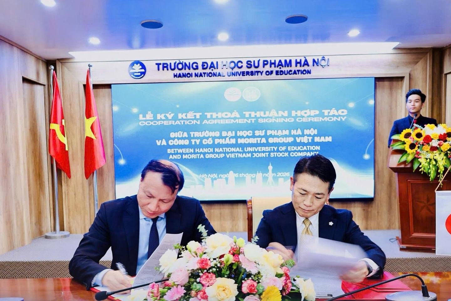 Hiệu trưởng Trường Đại học Sư phạm Hà Nội và Tổng Giám đốc Morita Group Việt Nam ký Biên bản ghi nhớ hợp tác, phía sau là màn hình Lễ ký kết thỏa thuận hợp tác