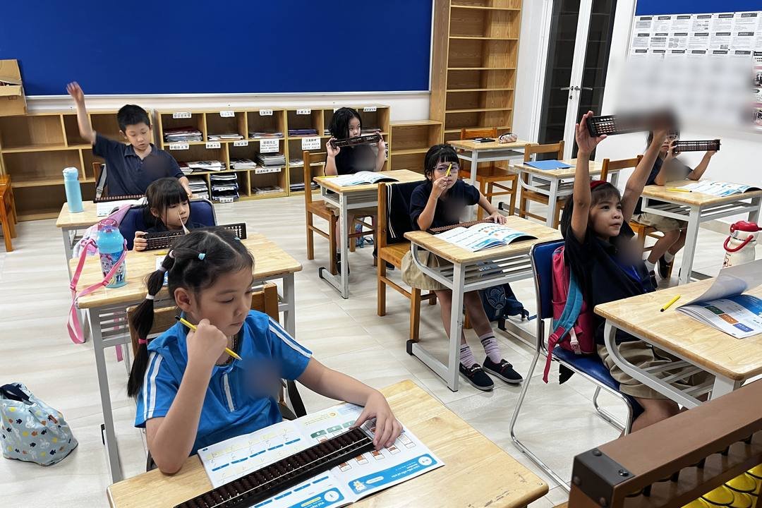 Bé gái mặc áo xanh nhạt ngồi ở bàn đầu, tay cầm bút chì đưa lên cằm, mắt nhìn quyển sách đang mở; trên sách là bàn tính Soroban; phía sau, nhiều bạn giơ bàn tính lên cao