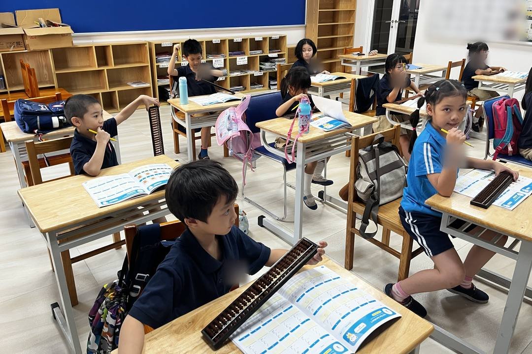 Lớp học Soroban: các bạn ngồi ở bàn riêng, trên bàn có bàn tính và sách đang mở; cậu bé ngồi gần nhất đặt tay lên bàn tính và nhìn lên phía trước