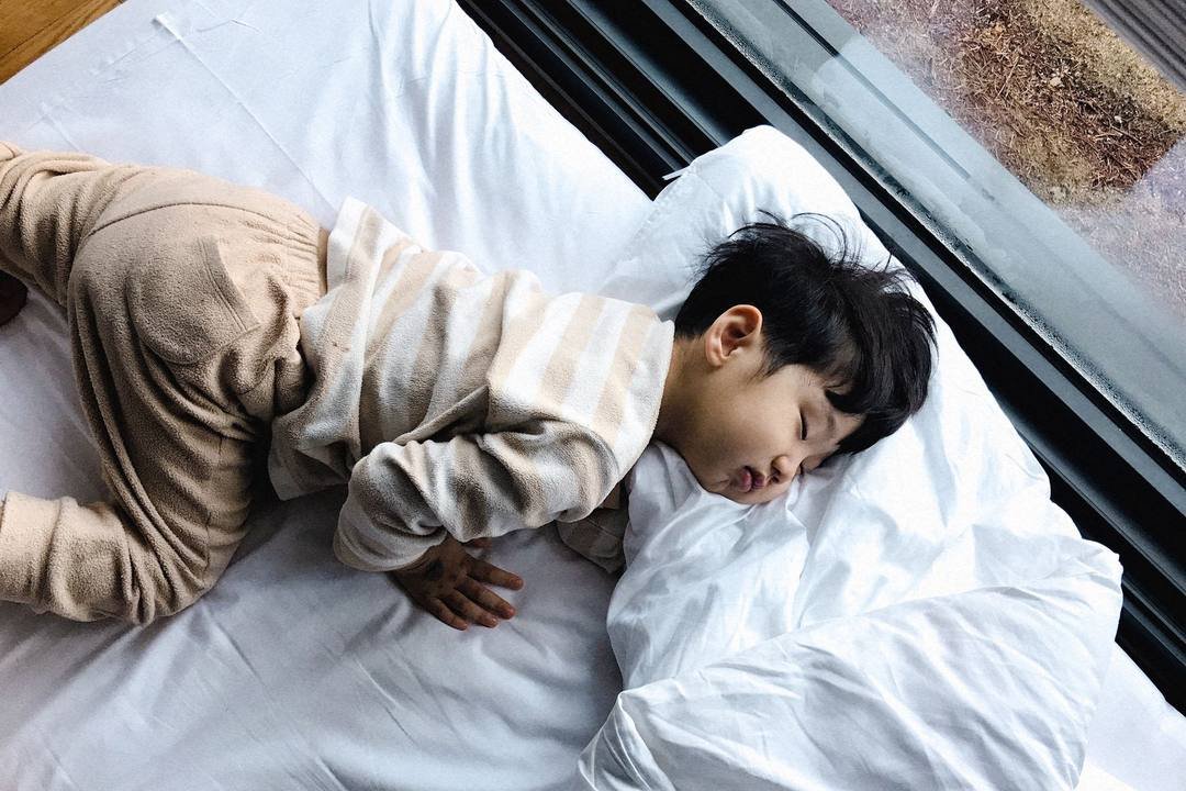 Bé trai tóc đen ngủ say trên giường trắng cạnh cửa kính, chăn gối hơi xộc xệch