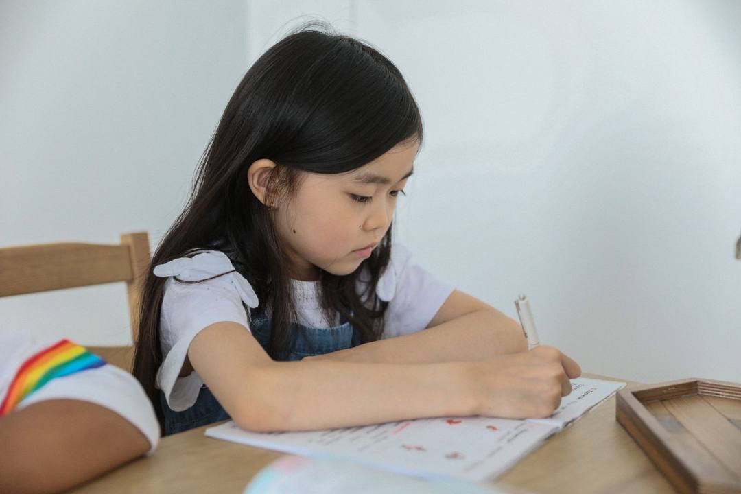 Bé gái tóc dài ngồi bên bàn gỗ, cúi xuống cầm bút viết vào quyển vở bài tập đang mở