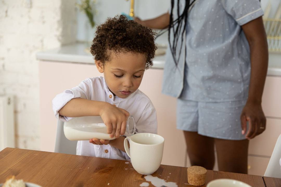 Bé trai tự cầm chai rót sữa vào cốc, trên bàn gỗ có vài vệt sữa bị đổ, một người lớn đứng ngay bên cạnh