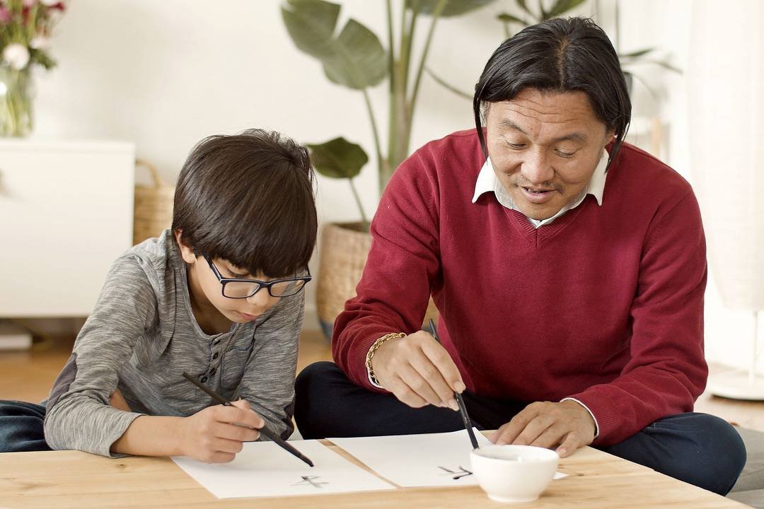 Một người đàn ông và một bé trai đeo kính ngồi bên bàn thấp, cùng cầm bút lông viết chữ lên giấy trắng