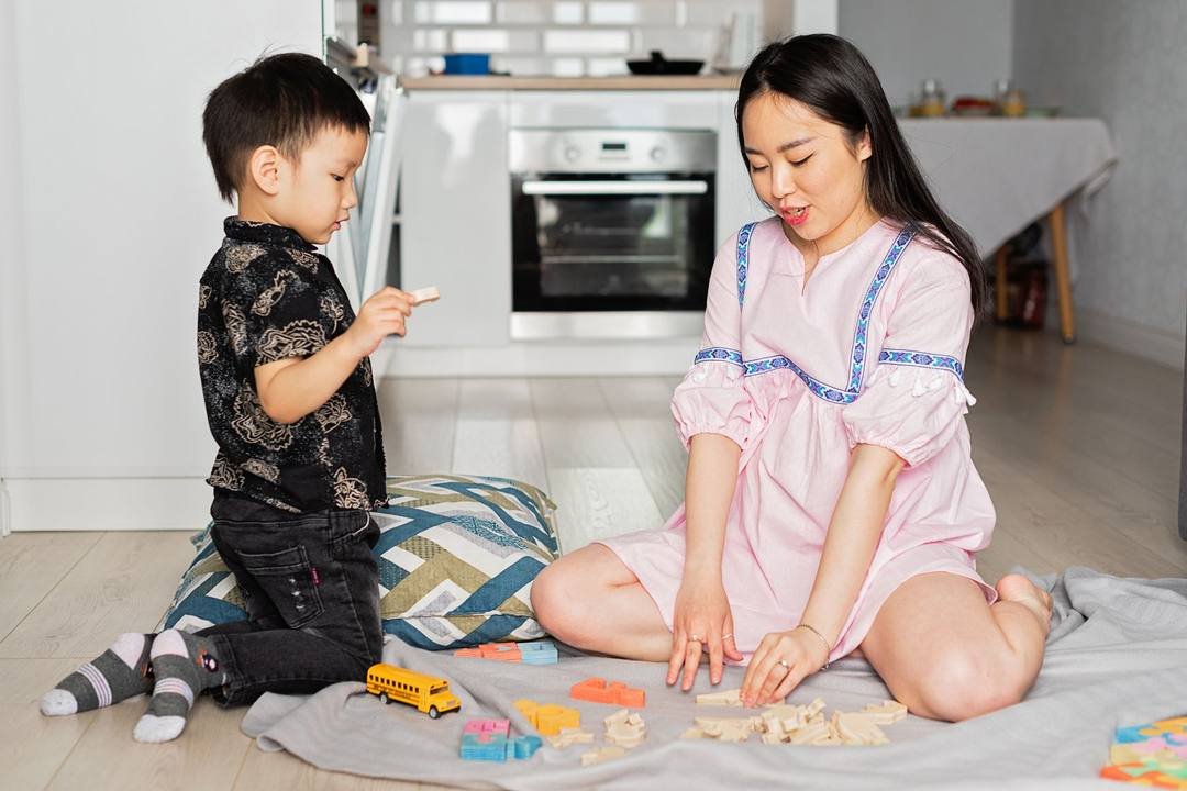 Mẹ và bé trai ngồi trên sàn gần gian bếp, chơi với các miếng gỗ, khối nhựa nhiều màu và một chiếc xe buýt đồ chơi màu vàng