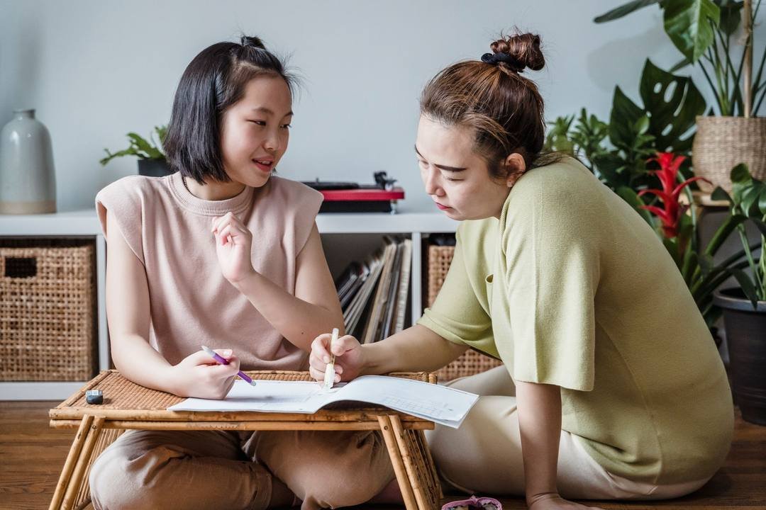 Mẹ và con gái ngồi trên sàn bên chiếc bàn mây thấp; mẹ cầm bút chỉ vào trang vở đang mở, con gái cầm bút màu tím, vừa nói vừa nhìn mẹ; phía sau là kệ có giỏ mây và nhiều chậu cây xanh