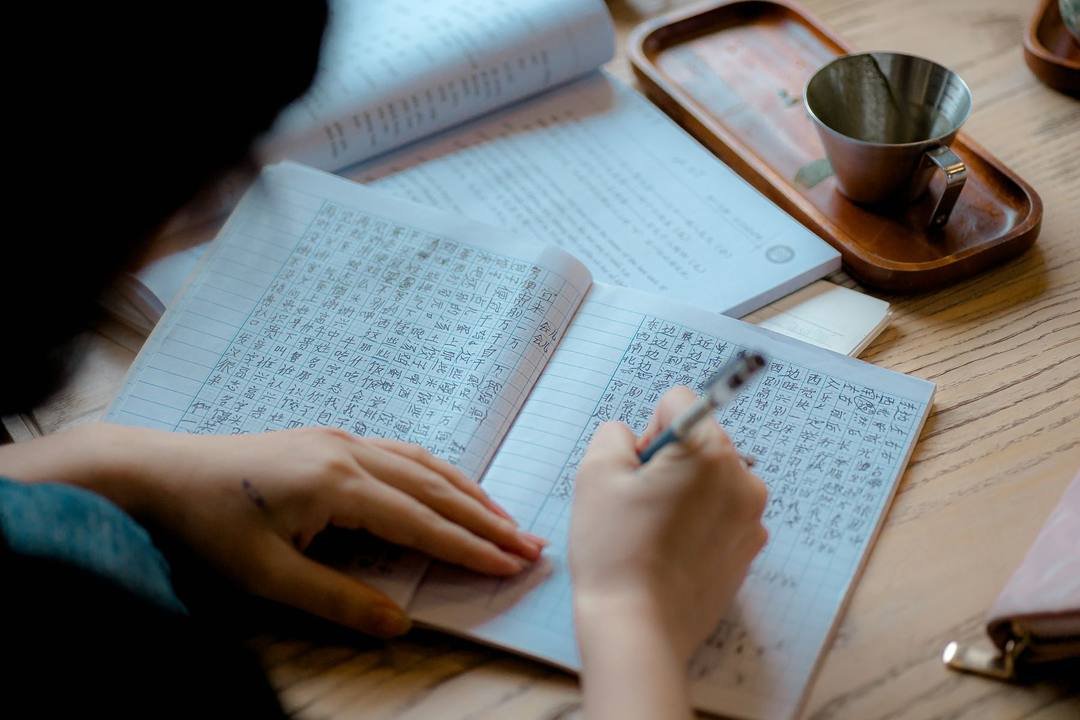 Bàn tay cầm bút viết chữ vào cuốn vở kẻ ô vuông nhỏ, trên bàn gỗ có sách và khay trà