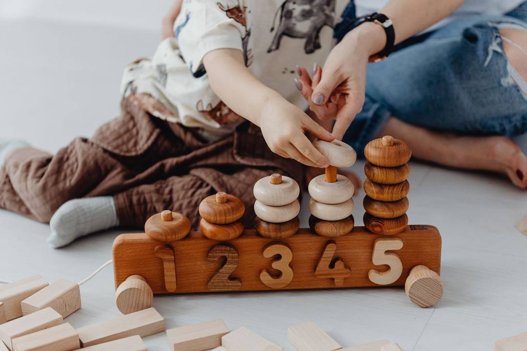 Bàn tay em bé đặt một chiếc vòng gỗ vào cọc trên giá gỗ có các số 1, 2, 3, 4, 5; bên cạnh là tay người lớn, dưới sàn có các khối gỗ